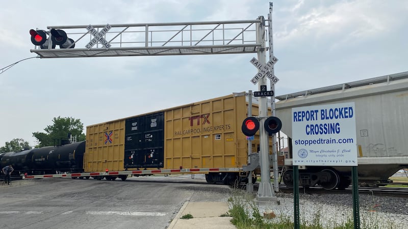 A freight car blocks a street and sidewalk, and the lights on the crossing warning are...