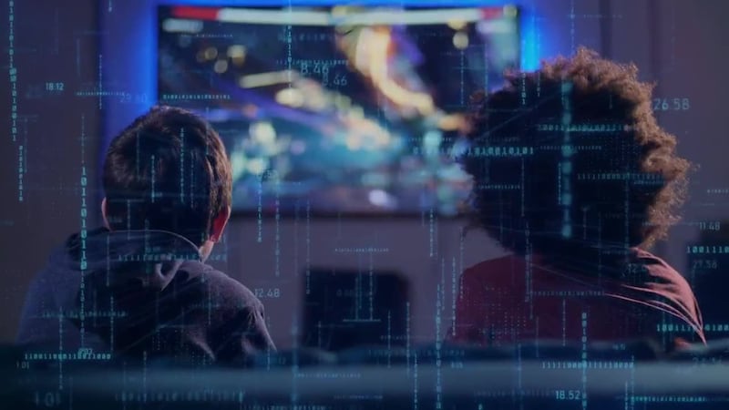 A photograph of two teens seated on a couch, seen from behind looking at a television screen....